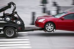 picture of the rear end of a two wheel lift tow truck towing a small red car quickly on a city street