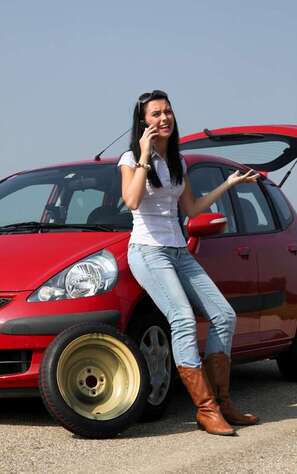 a small red hatch back car with a flat tire and the back hatch open with a long brown haired wearing a white t-shirt jeans and cowboy boots as she talks on the phone in front of car with left hand raised 
