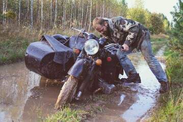 man waering camo jacket trying to push vintage black motorcycle with side car suck in flooded muddy dirt road