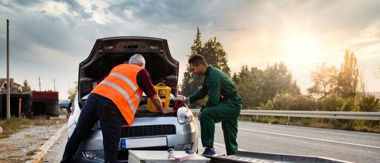 a cloudy day with the sun setting to the right as 2 tow truck drivers provide roadside assistance to a white vehicle with its hood up for a jump start. The wheel ramps of the flatbed tow truck are visible in the foregroundd